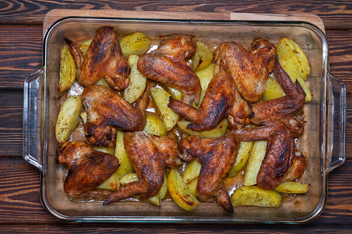 Baking sheet with baked chicken wings with spices and potatoes on a wooden table, top view